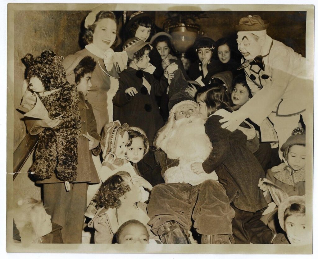 1950s vintage Christmas Photo: Santa is surrounded by loads of kids and one creepy clown, while clearly over the whole experience.