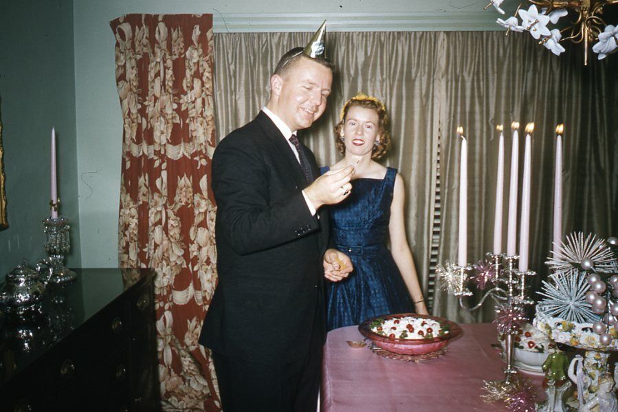 1950s vintage photo of a couple in 1950s Fashions celebrating New Years Eve with a tiny party hat and table filled with fun 1950s party foods. 