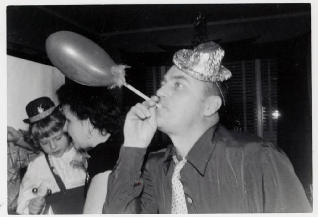 1950s vintage photo of a man in a party hat blowing on a balloon at a New years eve party in 1955. 