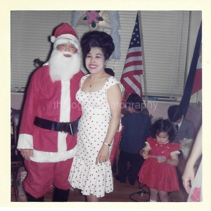 1960s vintage photo of a pretty wman in a polka dot 1960s dress and big 60's hairstyle posing with a man dressed up as Santa at a Christmas party with kids in the background. 