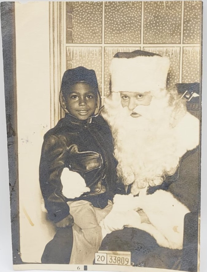 vintage photo from the mid century of a young Black boy in a leather jacket and leather hat sitting on Santas lap