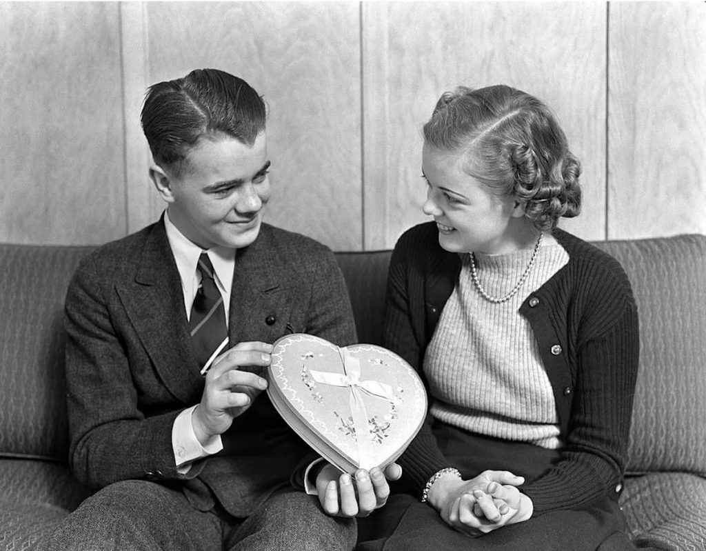 1930s vintage photo of two teenagers sitting on a couch together on Valentines Day. The young man is giving a box of chocolates to the young woman with the 1930s hairstyle