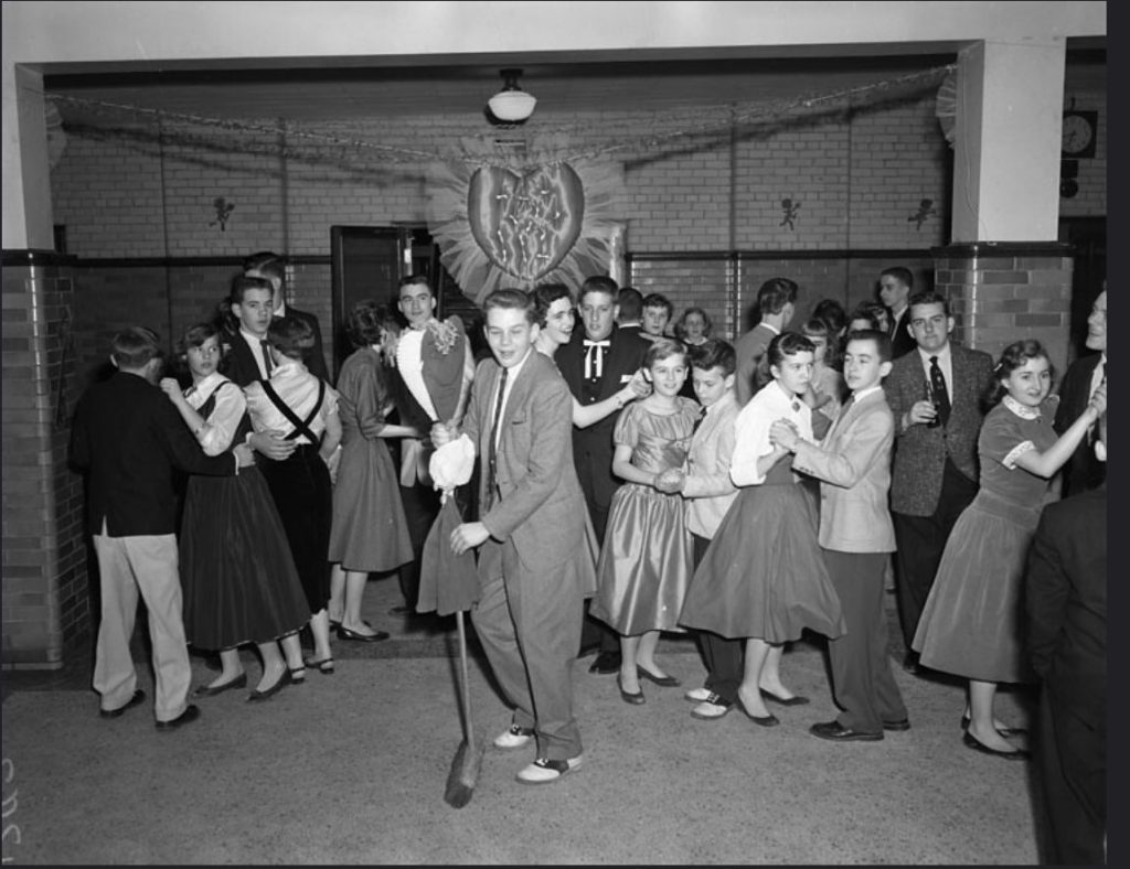 1950s vintage photo of a Valentine's Day Dance featuring young teenagers and a young man dancing with a broom made to look like his date. 