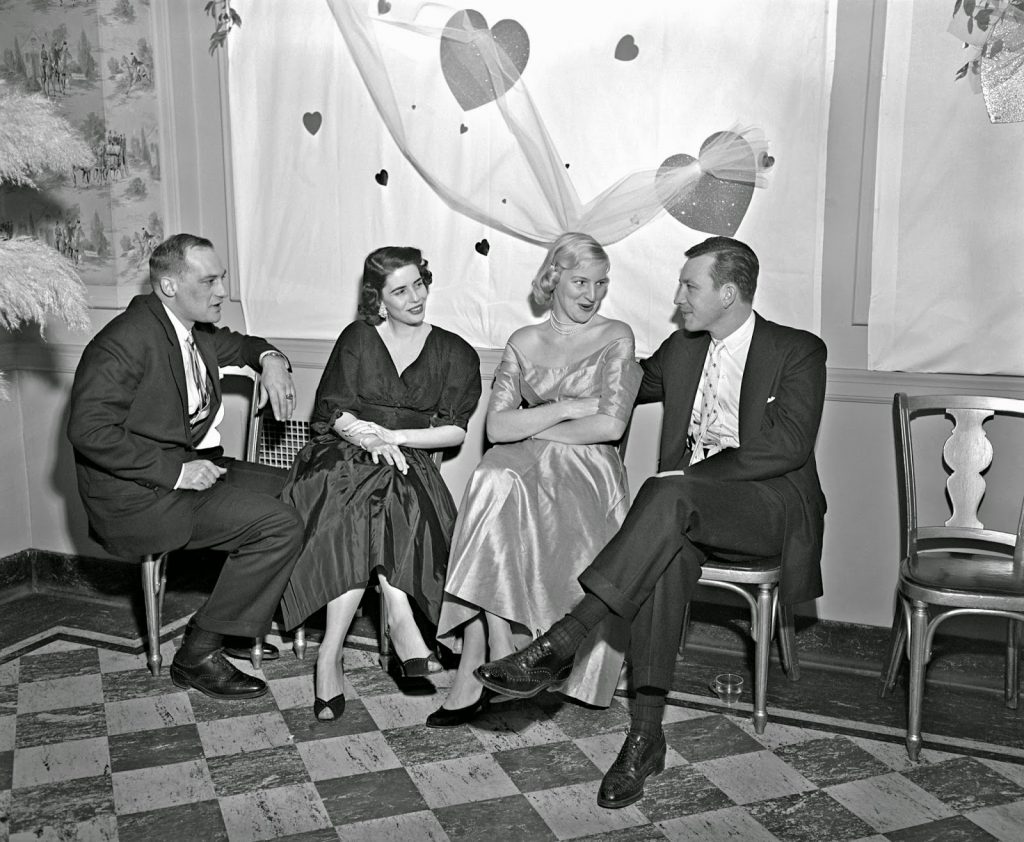 1950s vintage photo of a 1950s Valentines Day Dance featuring 2 couples sitting together in 1950s fashions with Valentine decorations on the wall. 