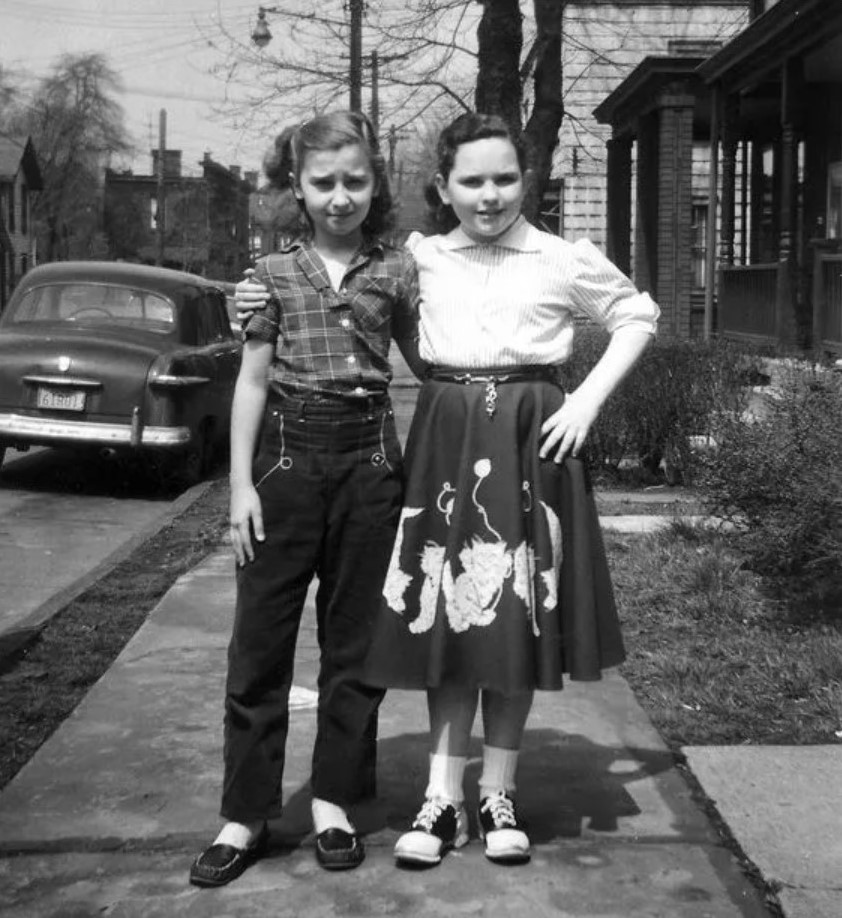 1950s vintage photo of two young girls in 1950s fashions. One girl is wearing jeans and plaid shirt and the other is wearing a felt circle skirt with an applique on it and saddle shoes.