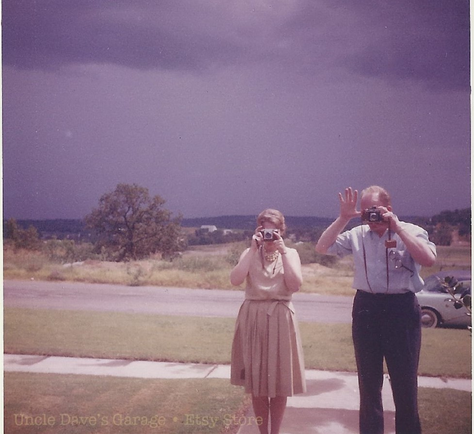 Say Cheese! 1960s vintage photo of a couple taking pictures with their camera's while on vacation with dark skies behind them. 