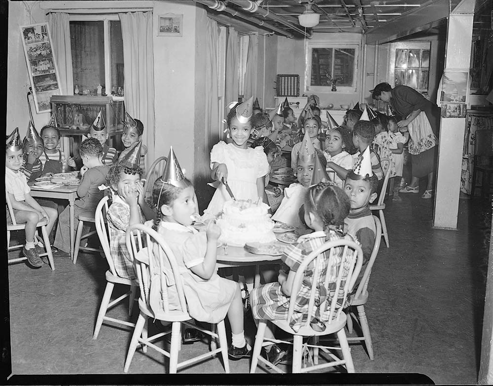 Late 1940s vintage photo of a group of young Black kids celebrating a birthday party at a nursery school. They are all wearing party hats and waiting for the birthday girl to cut the cake. 