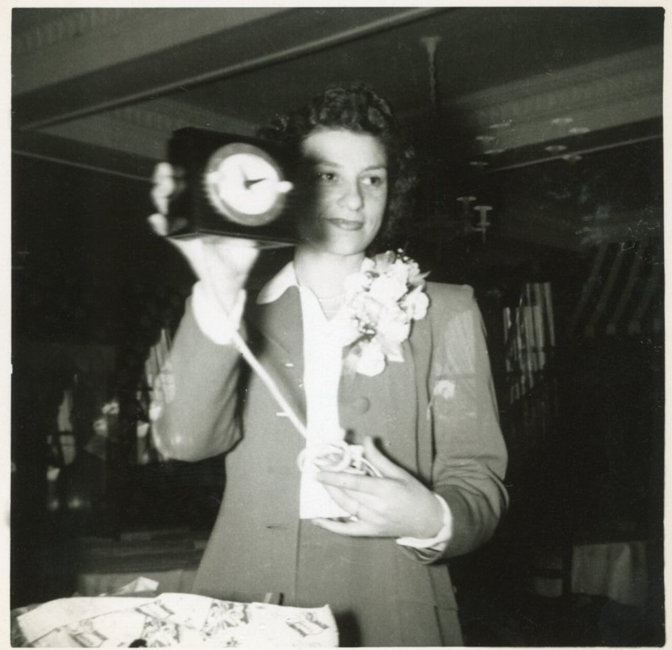 1940s vintage photo of a young woman at her bridal shower holding a gift of an electric alarm clock while wearing a large corsage.