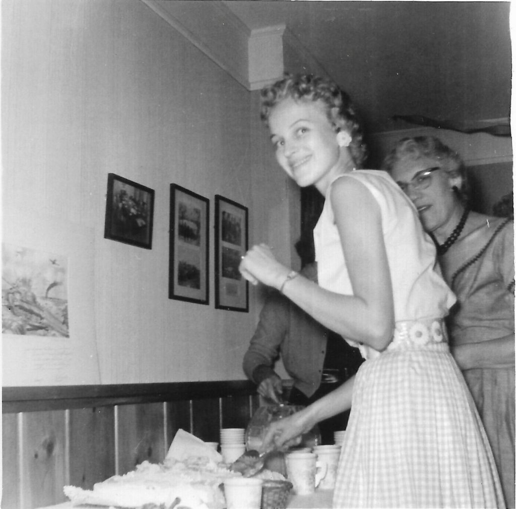 1950s vintage photo of a 1955 Bridal Shower featuring the bride to be posing while cutting the cake in a 1950s top and 1950s skirt. 