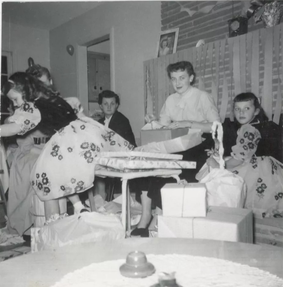 1950s vintage photo of a Bridal Shower featuring a pretty young woman opening her gifts with twin girls in matching dresses taking up the photo and streamers as decor from the shelf. 