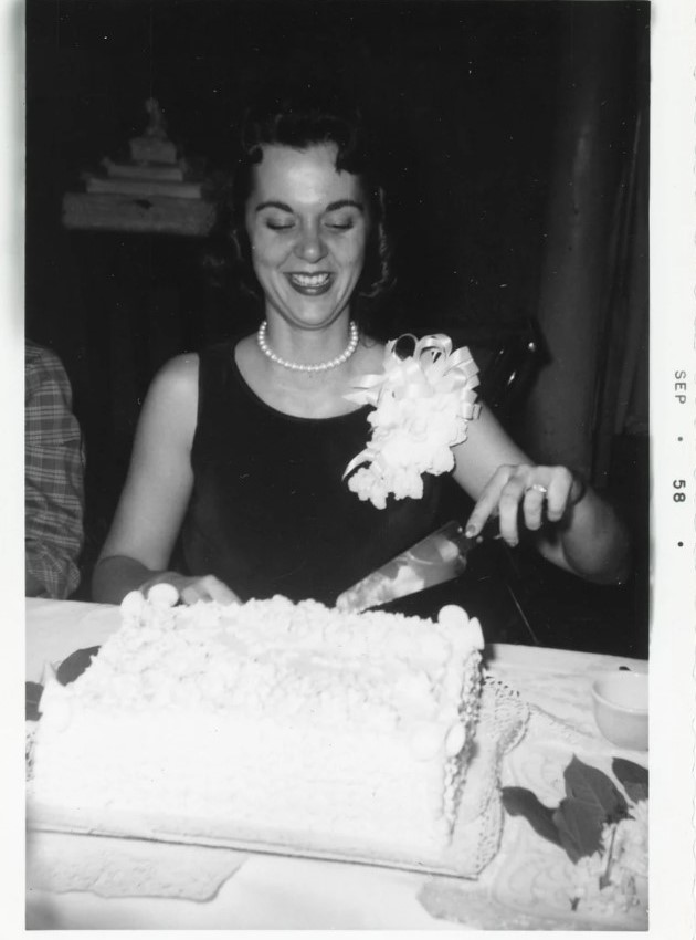 1950s vintage photo of a September 1958 vintage bridal shower. The young woman is wearing a large corsage and cutting her bridal shower cake.  