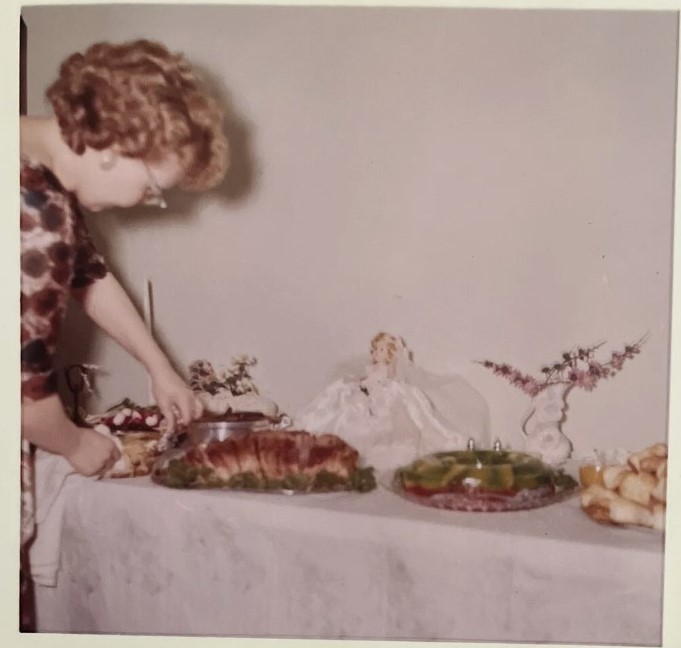 1960s bridal shower food table featuring a jello mold and a bride doll and a woman in a 1960s dress putting out food.