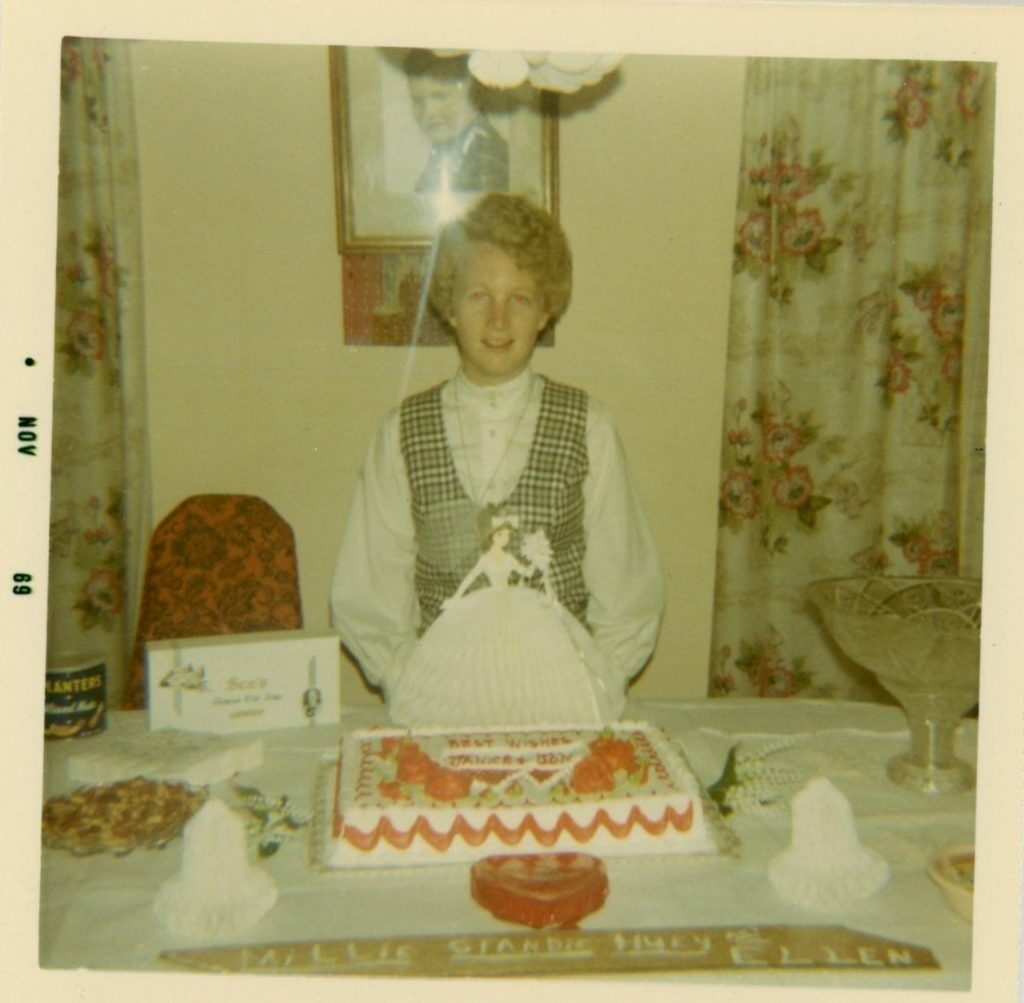 1960s vintage photo from November 1969 of a curly haired bride (Janice) posing with her cake and a cute bride doll in front of a family portrait and floral curtains at her bridal shower.