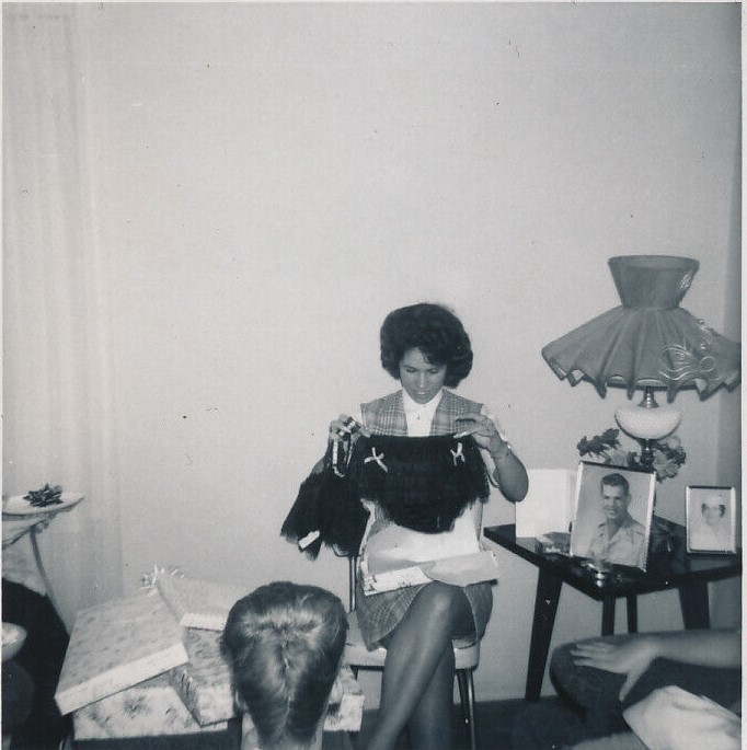 1960s vintage photo of a bridal shower from 1964 featuring the bride to be opening up lacy underwear while a friend with a big 1960s hairstyle looks on. The photo of the groom is on the table. 