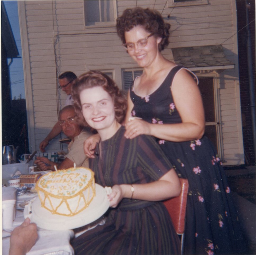 1960s vintage photo of a woman holding a heart shaped birthday cake with candles it while in a 1960s dress with her mom standing behind her also in a pretty 1960s dress. 