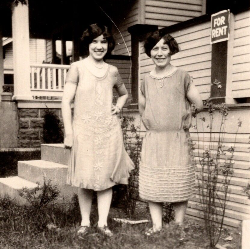 1920s vintage photo of a mother and daughter in 1920s dresses posing out of their home that says for rent. Fantastic 1920s hairstyle inspiration as well.