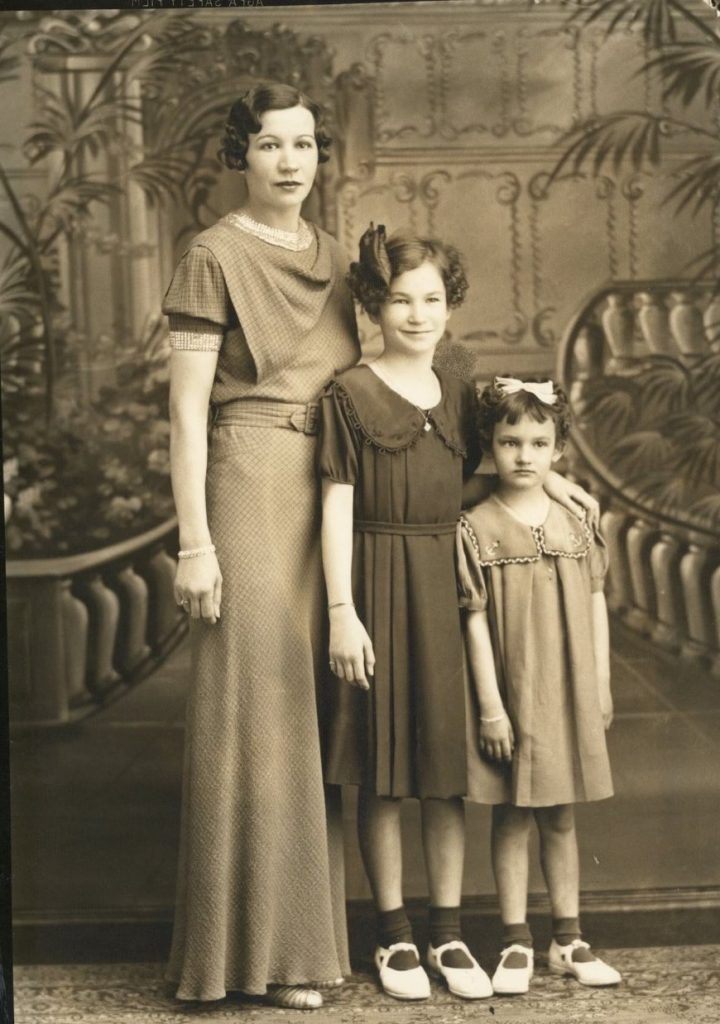 1930s vintage photo of a mother in a 1930s dress and early 1930s hairstyle posing with her cute daughters with bows in their hair and dresses for a studio portrait. 