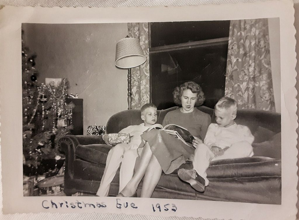 1950s vintage photo of Christmas Eve 1953 featuring an image of a mother on the couch with her kids in pajamas reading a book with presents by the Christmas Tree