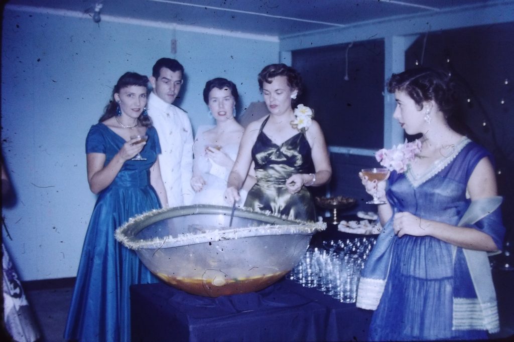 1950s vintage photo of a 1950s party with a large punch bowl in the middle while women in 1950s evening gowns and 1950s hairstyles stand around the bowl.