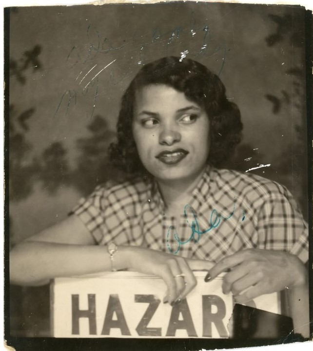 1950s vintage photo of a Black woman with a 1950s hairstyle and plaid dress on posing in front of a sign that says hazard