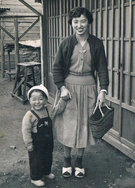 1950s vintage photo of a young Japanese mother in a 1950s skirt and cardigan holding the hand of her cute song in overalls and hat. 