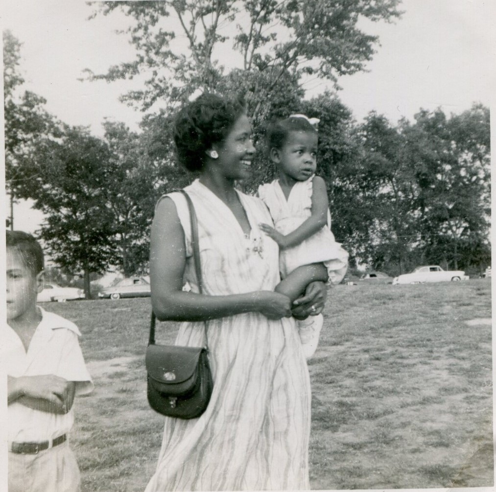 1950s vintage photo of a young Black mother in a simple 1950s dress holding her cute little girl in her arms while a young boy stands by