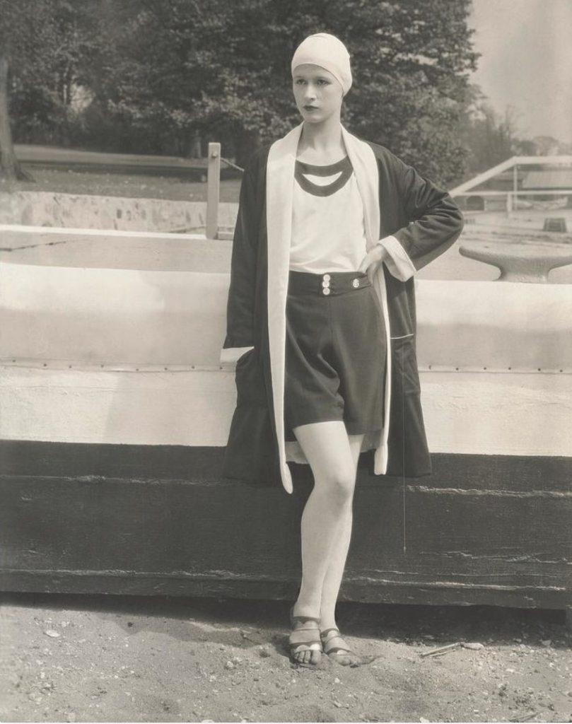 1920s Vintage Photo / 1920s Fashion: Vogue, 1927. Model, Portia Grafton, standing beside the gunwale of a boat in Great Neck, Long Island, wearing a black crepe beach coat with white terry lining and matching black shorts by Jean Patou, with a white sleeveless top by Kurzman, and a fitted white cap.