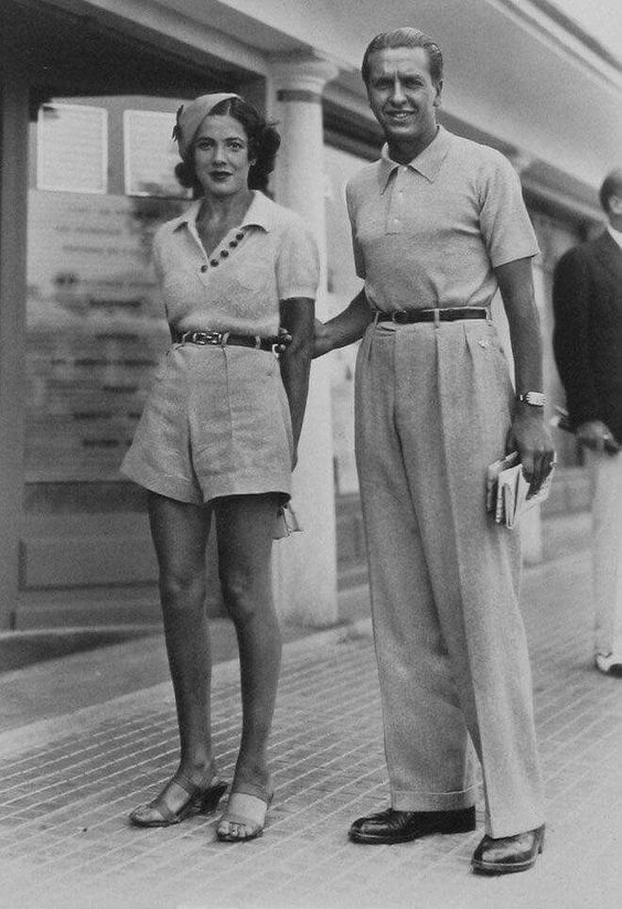 1930s vintage photo of a woman and man in 1930s fashions in Paris. The woman is wearing shorts, knit top and a hat and sandals. Our man is wearing pants and a golf shirt. 