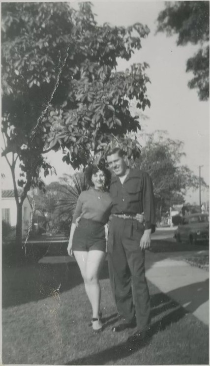 1940s vintage photo of a couple posing outside together. The woman is wearing shorts and 3 quarter sleeve top. The man appears to be in a working uniform for maybe a mechanic. 