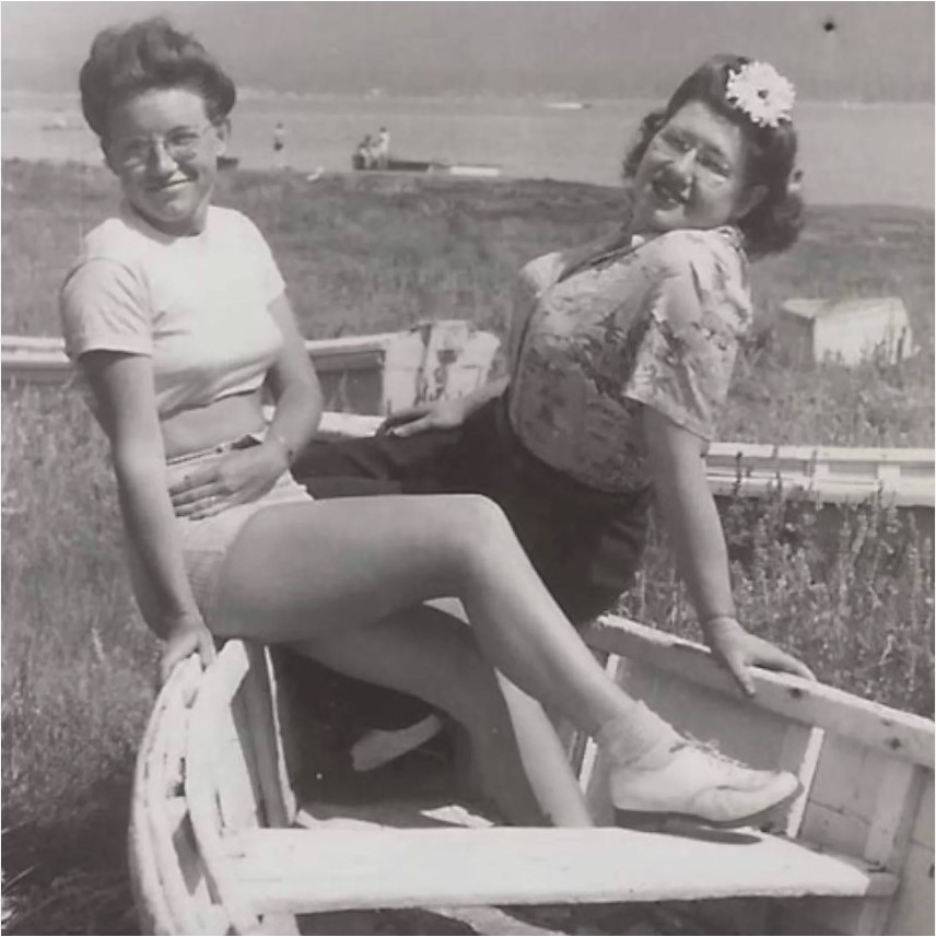 1940s vintage photo of two young women hanging out at the seaside in 1940s summer outfits and eyeglasses and 1940s hairstyles with a hair flower