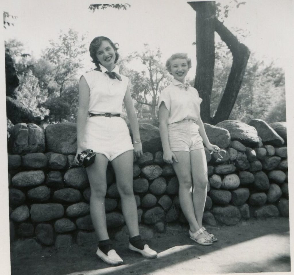 1950s vintage photo of 2 young women in shorts and blouses with scarf around their neck sightseeing as one girl is holding a camera. Cute 1950s Summer Fashion Inspiration!