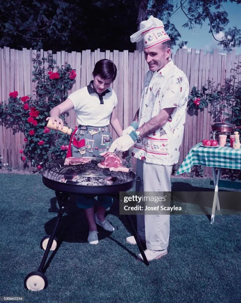 1950s vintage photo of a fatgher and daughter cooking steaks together in cute 1950s summer fashions and the dad is wearing a chefs hat