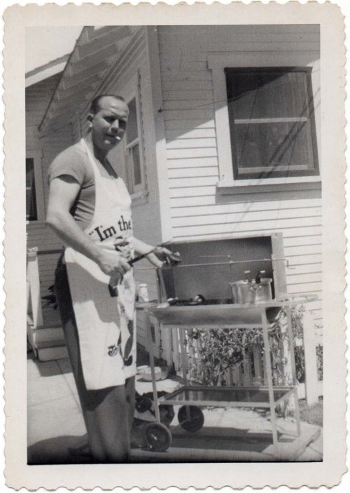 1950s vintage photo of a man having a BBQ in his backyard while wearing a chef apron and smoking a cigarette