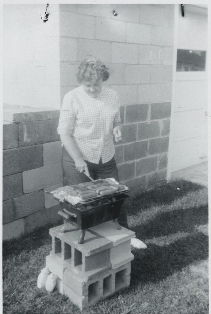 1950s vintage photo of a woman having a barbeque and flipping sausages outside of her house.