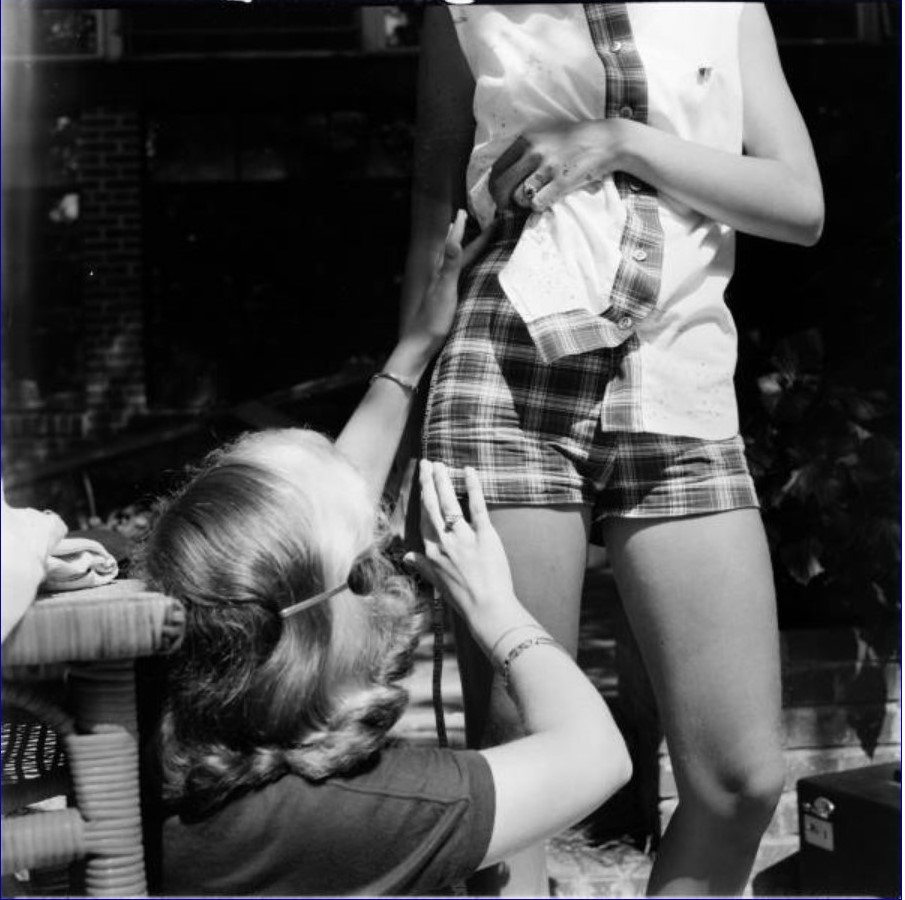 1950s vintage photo of a model getting her shorts fixed for a photo shoot about SHORT SHORTS. A woman with a 1950s hairstyle is helping her. 