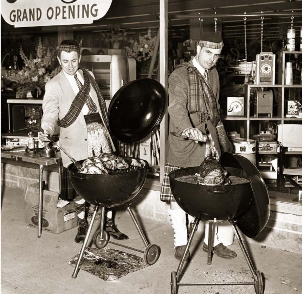 1950s vintage photo of two men in Scottish kilts grilling on their BBQ outside of a store opening celebration.