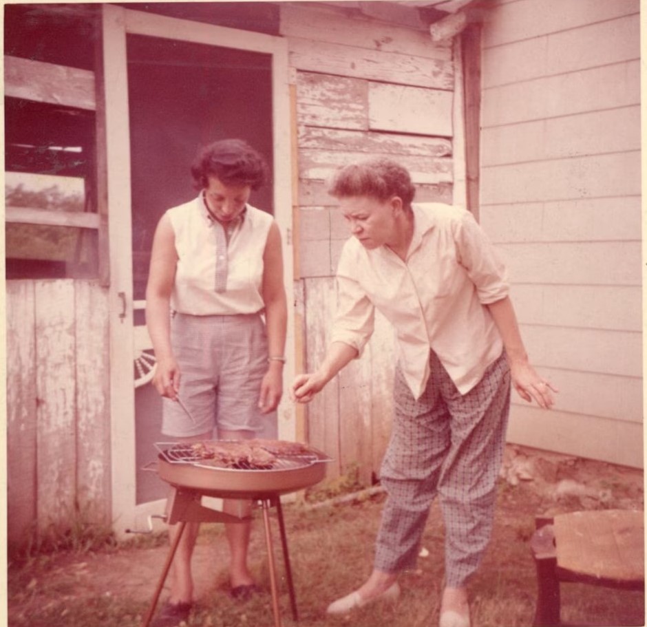 1960s photo of two ladies very focused on cooking the meat on their little grill in their backyard while wearing summer fashions like shorts. 