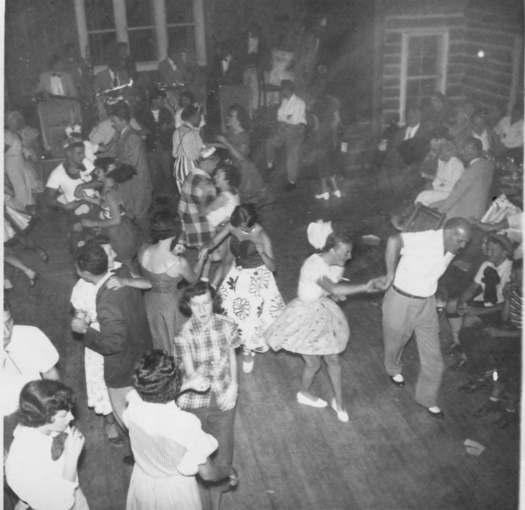 1950s vintage photo of couples dancing to a live band outside in 1950s fashions and 1950s hairstyles. Looks like some are dancing Jive.