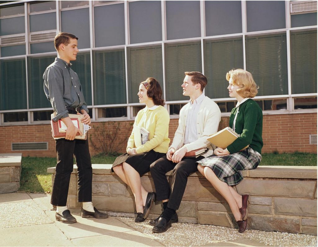 1960s vintage photo of high school students in 1960s fashions for fall sitting outside of their school chatting. 