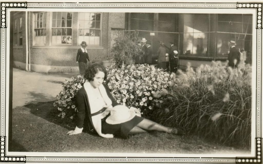 1930s photo of a young woman named Clara Foster with fingers waves in her hair and a long cardigan, poses casually in a random flower garden. 