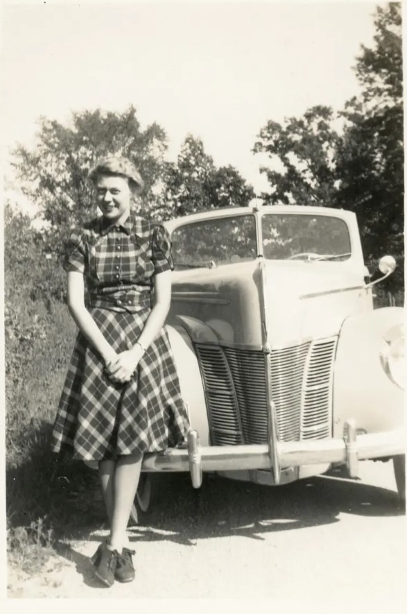 1940s vintage photo of a young woman in a plaid dress posing in front of a car. 