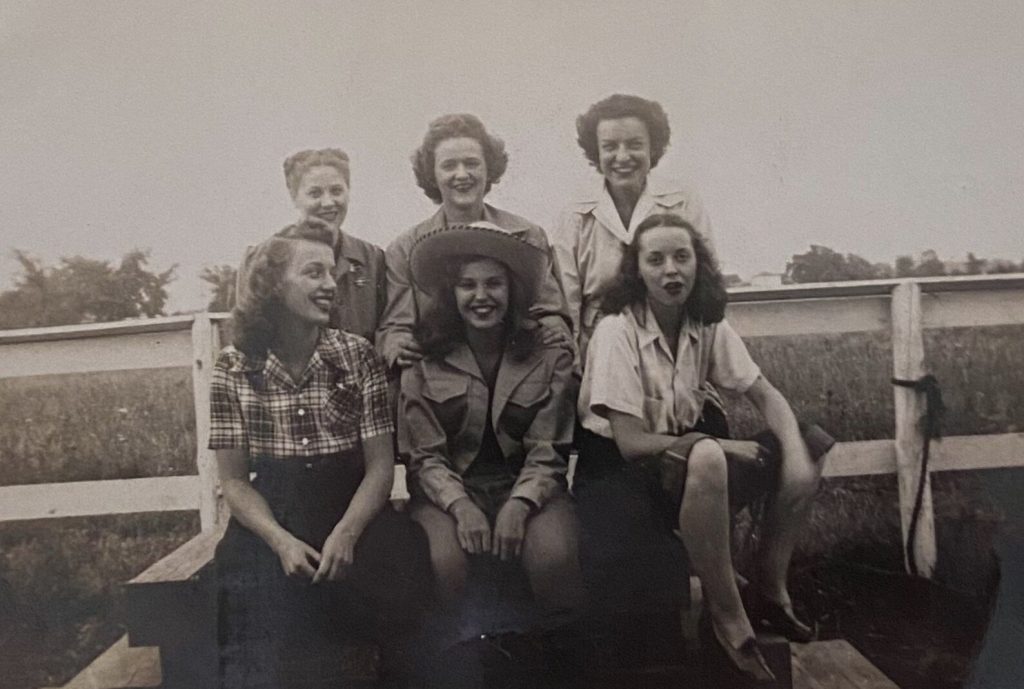 1940s vintage photo: Vintage Image of a group of young women in 1940s fashions and 1940s hairstyles posing together on a farm. One girl is wearing a plaid shirt and another girl is wearing a cowboy hat. 