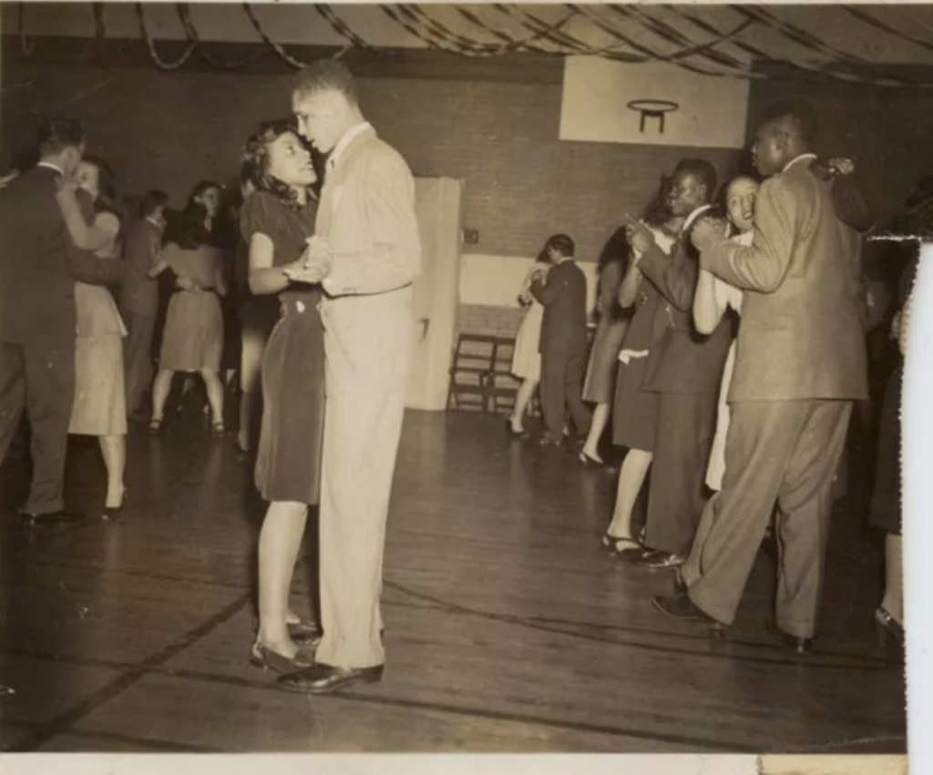 1940s vintage photo of a dance in a gym with young men and women. The photo features stylish Black Men and Women in the front of the photo. 