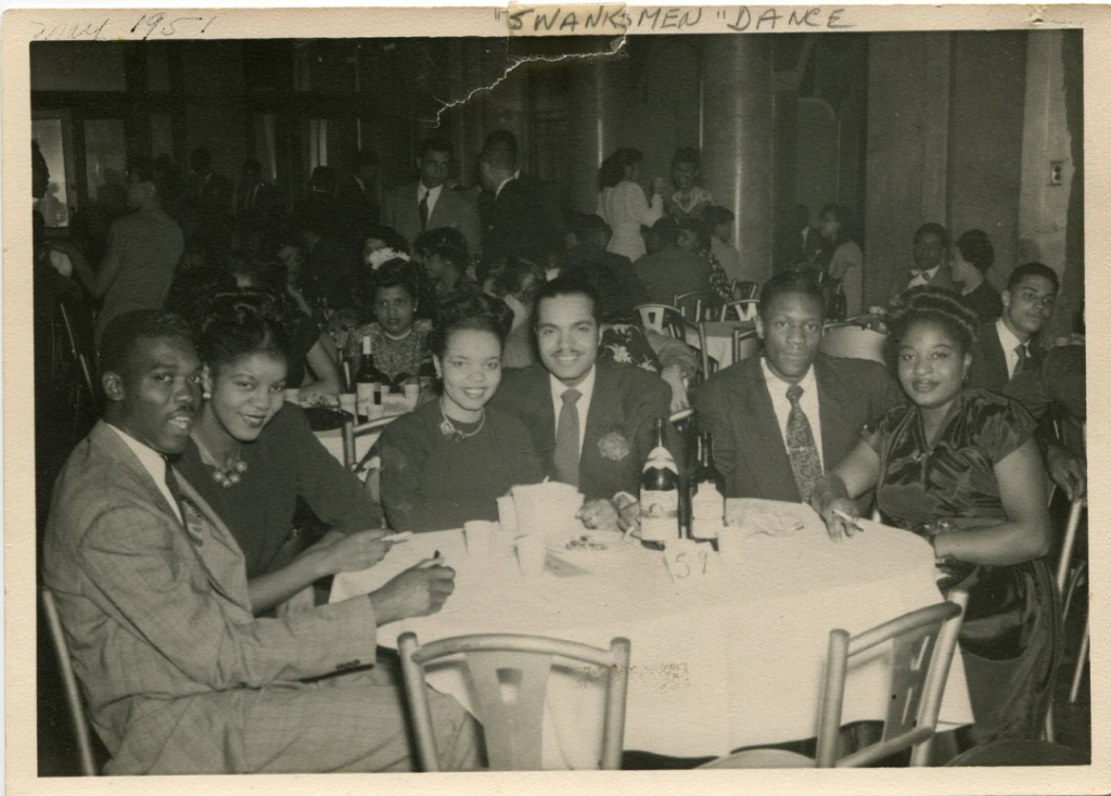 1950s vintage photo of a group of Black Men and Black Women all dressed up in 1950s fashions and 1950s hairstyles sitting at a table at a dance. 