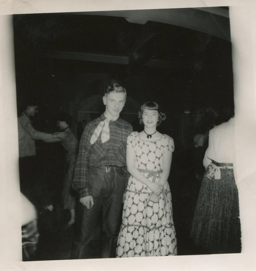 1950s vintage photo of a young couple at a dance wearing western wear fashions. Couples are dancing in the background. 