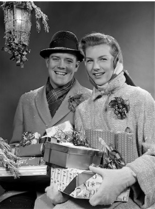 1950s Vintage Photo of a couple in 1950s Winter Coats holding Christmas Presents. The 1950s Woman is wearing a Christmas Corsage. 