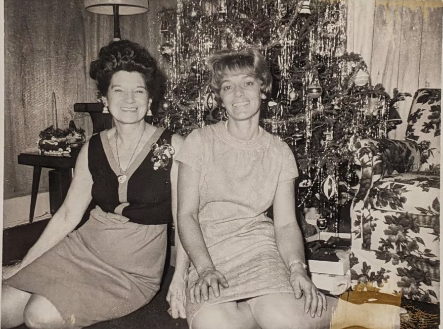 1960s Vintage Photo: Two Women in 1960s Fashions and 1960s hairstyles smile for the camera in front of a Christmas Tree. The one woman is wearing a Christmas Corsage. 