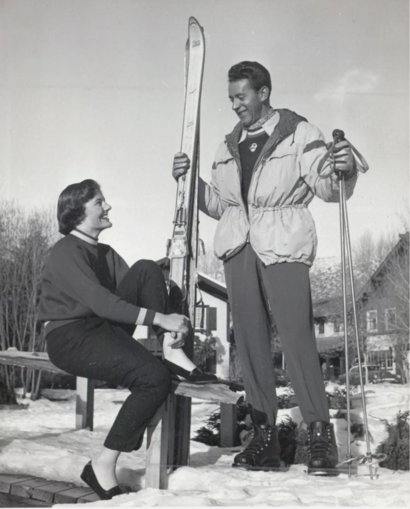 1950s vintage photo of a man in 1950s ski fashion talking to a woman in 1950s clothing at a ski resort. 